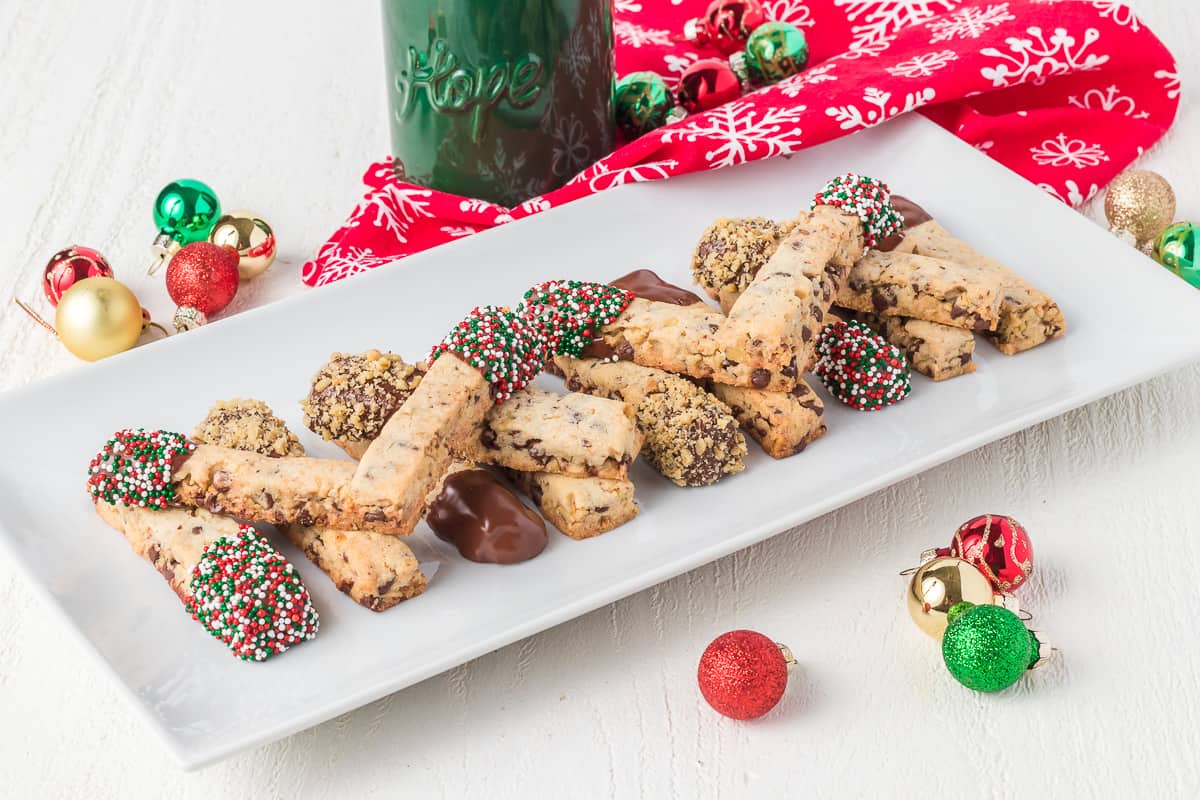 A white rectangular plate with biscotti, some dipped in chocolate and covered with sprinkles or nuts, surrounded by Christmas ornaments and a red holiday cloth.