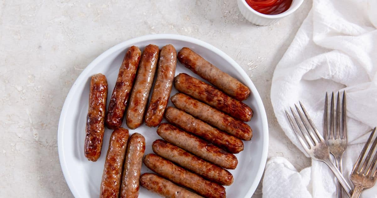 A white plate with cooked sausage links next to a small cup of ketchup, silver forks, and a white cloth napkin on a light surface.