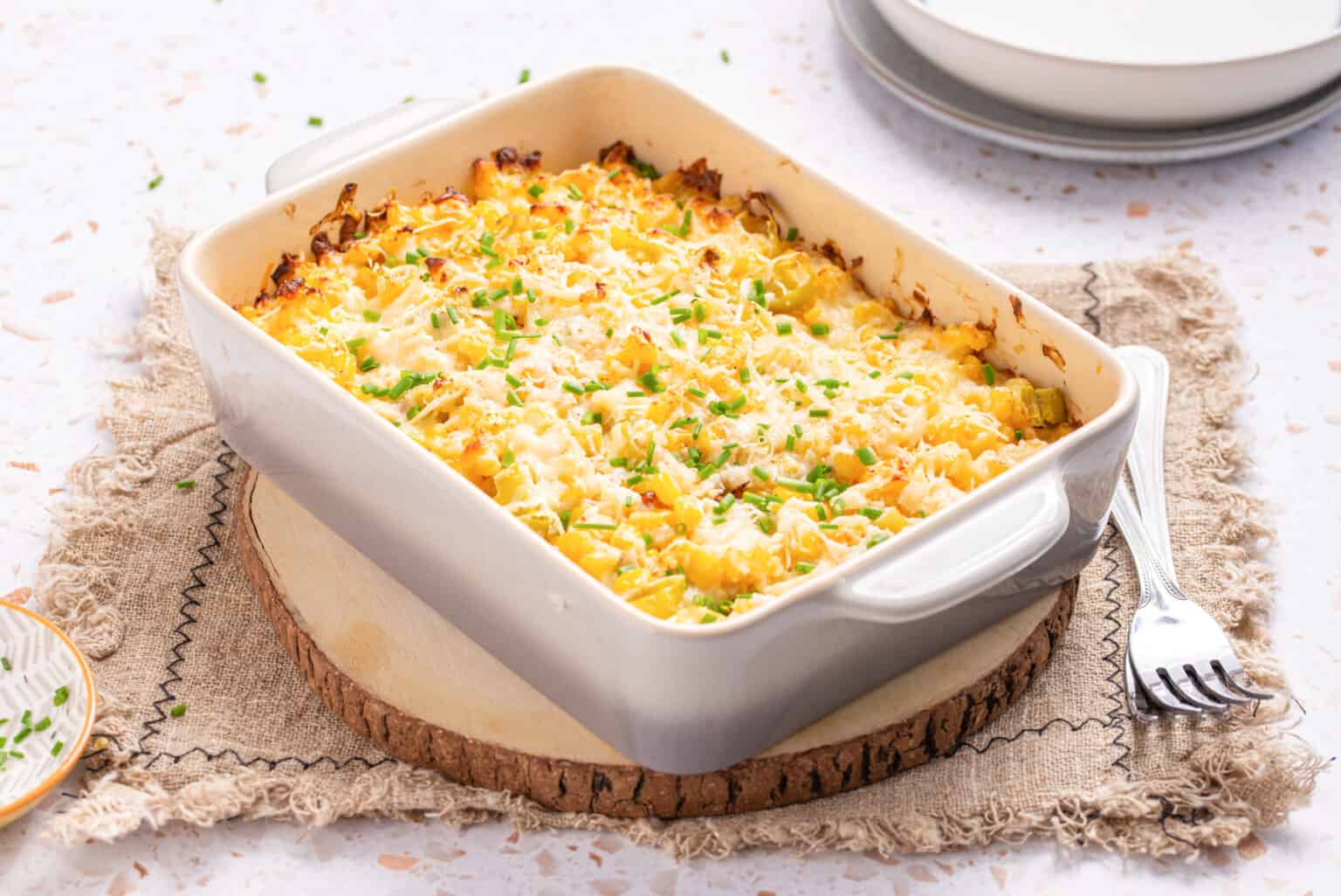 A baked casserole topped with melted cheese and chopped chives in a white dish, placed on a rustic cloth and wooden board, with plates and utensils nearby.