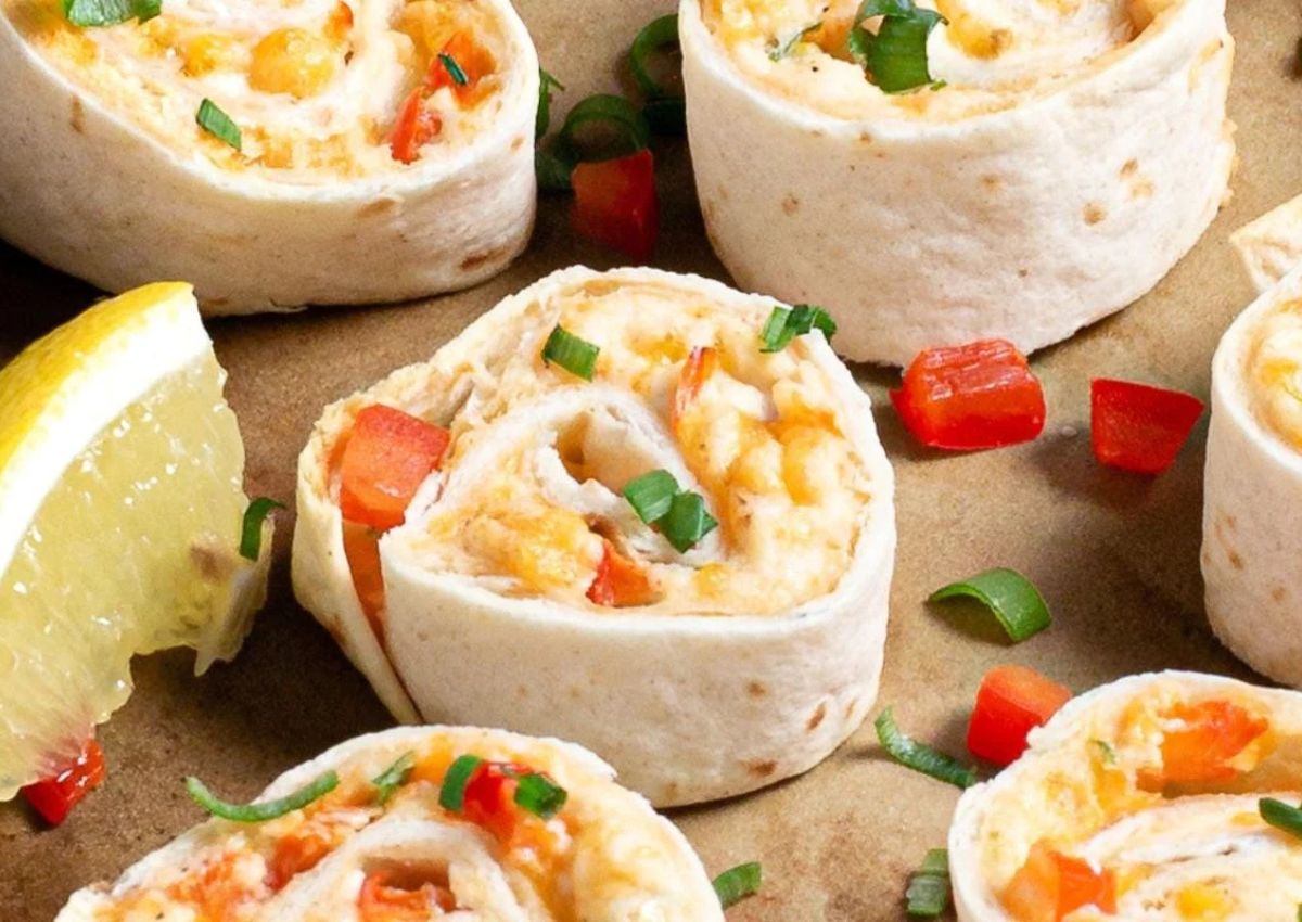 Close-up of tortilla pinwheels filled with cheese, diced red peppers, and green onions, arranged on a brown surface next to a lemon wedge.