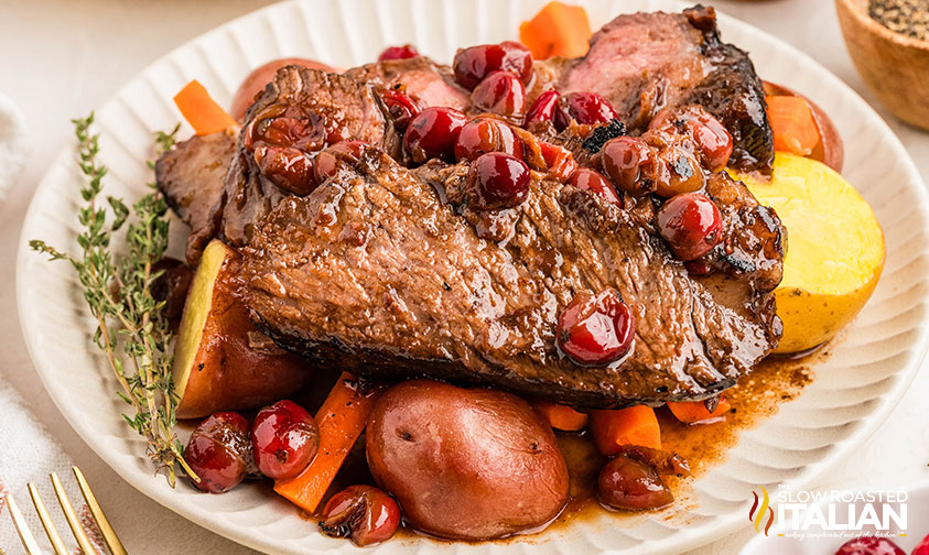 Slices of roast beef topped with cherries served on a plate with red potatoes, carrots, and sprigs of thyme.