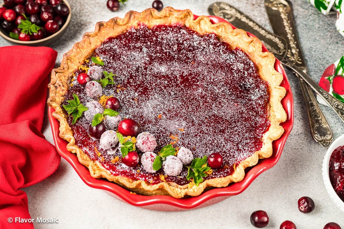 A cranberry pie in a red pie dish is topped with sugared cranberries, mint leaves, and powdered sugarโan inviting choice for Christmas Pie Recipes. Utensils and a festive red napkin are set nearby.