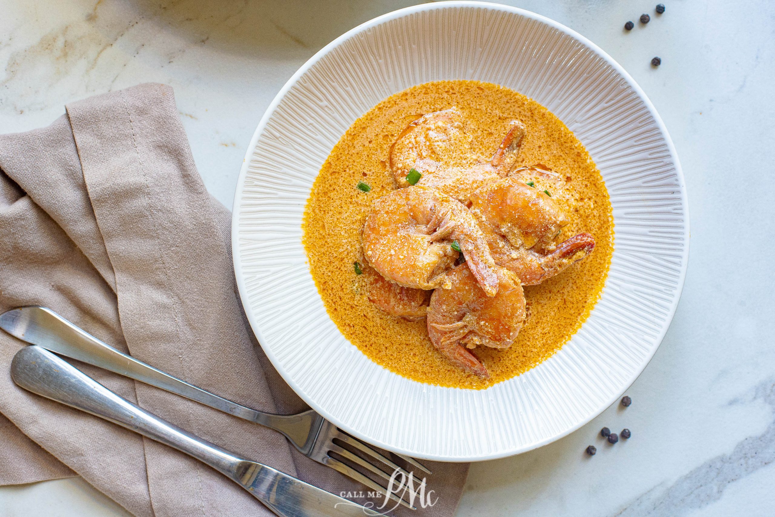 A white bowl filled with shrimp in creamy orange sauce, placed on a marble surface beside a folded beige napkin, fork, and knife.