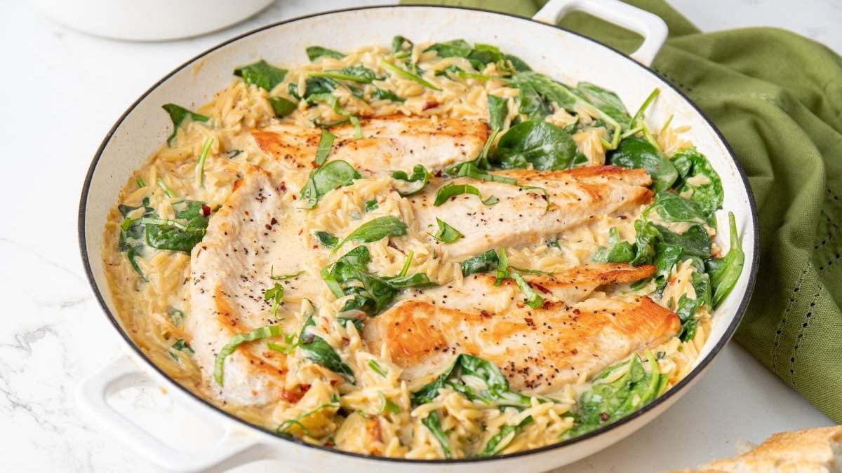 A skillet with seared chicken breasts, creamy orzo pasta, and spinach, all combined in a rich sauce.