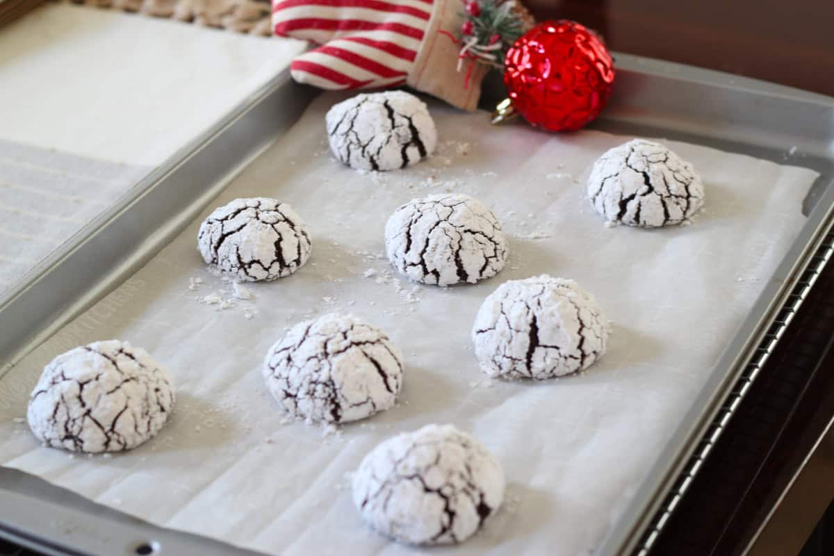 A baking tray with nine chocolate crinkle cookies dusted with powdered sugar on parchment paper; a red ornament and striped napkin are in the background.