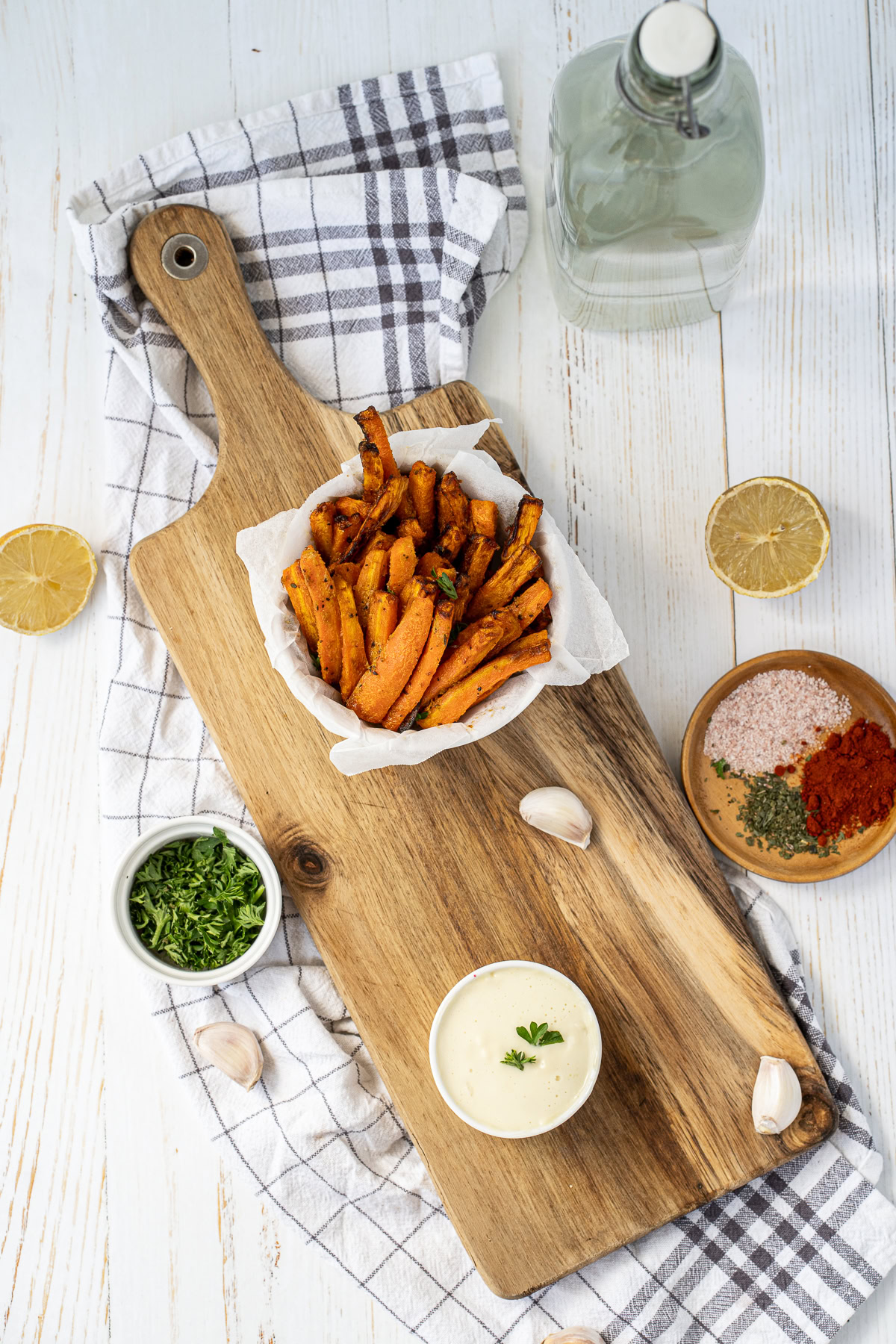 A serving of sweet potato fries in a paper-lined basket on a wooden board, with dipping sauce, herbs, spices, lemon slices, a glass bottle, and garlic cloves arranged around.