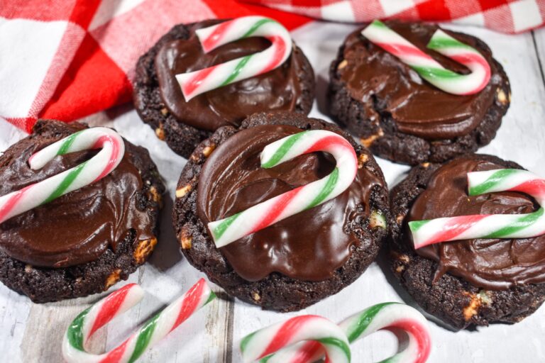 Five chocolate cookies topped with chocolate frosting and mini red, white, and green candy canes are arranged on a white wooden surface with a red and white checkered cloth.