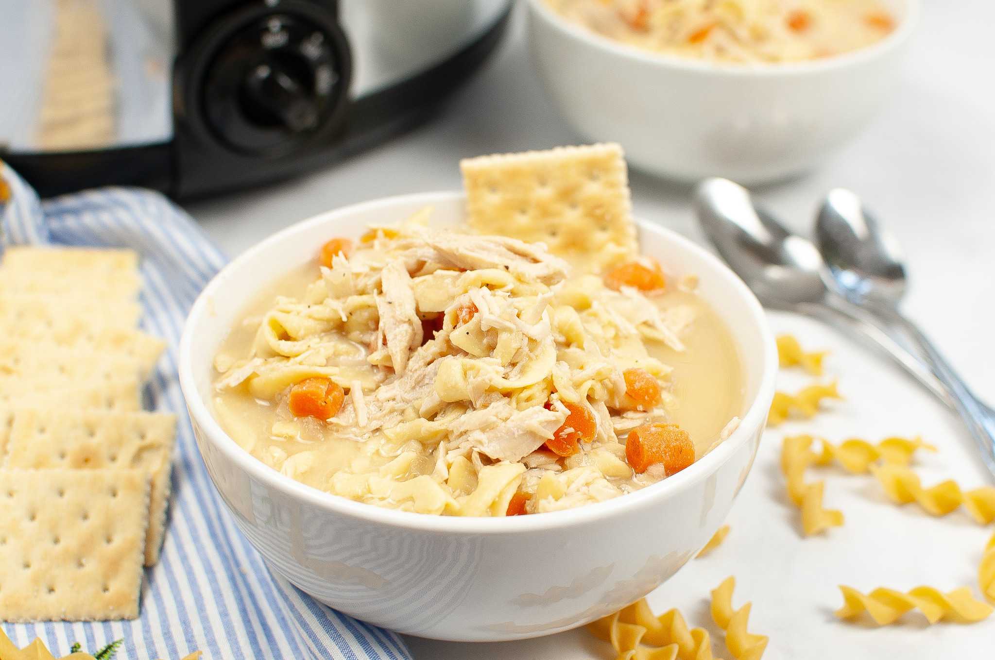 A bowl of chicken noodle soup with carrots and crackers, with a bowl of soup, spoons, and spiral noodles in the background.