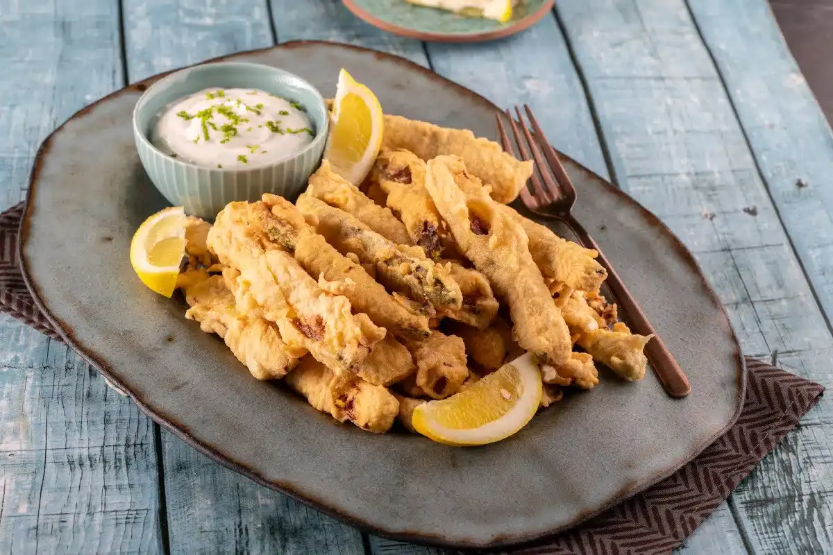 A plate of fried battered strips served with lemon wedges and a bowl of white dipping sauce, set on a rustic blue wooden table.