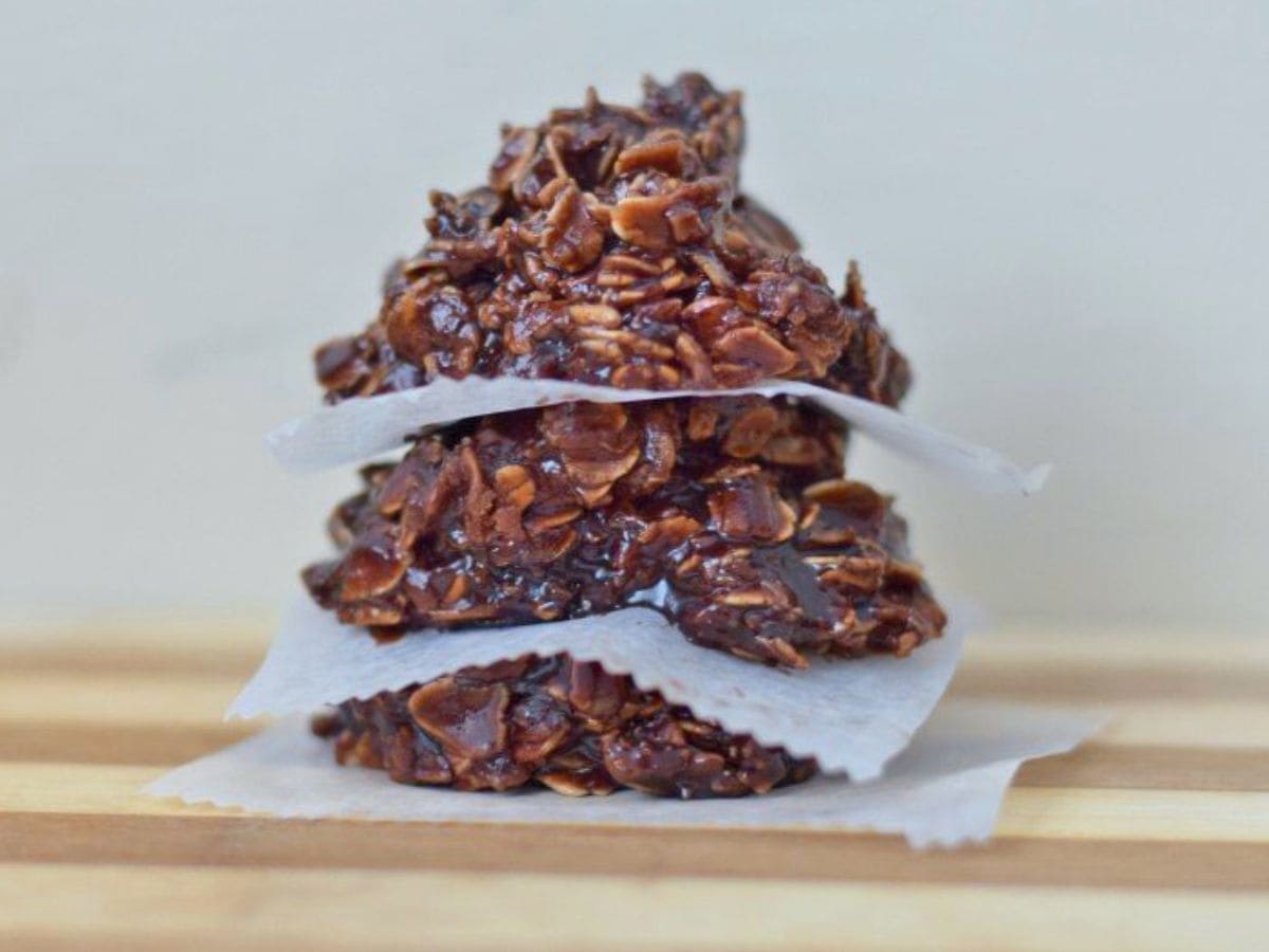A close-up of three no-bake chocolate oatmeal cookies stacked with pieces of parchment paper between them on a wooden surface.