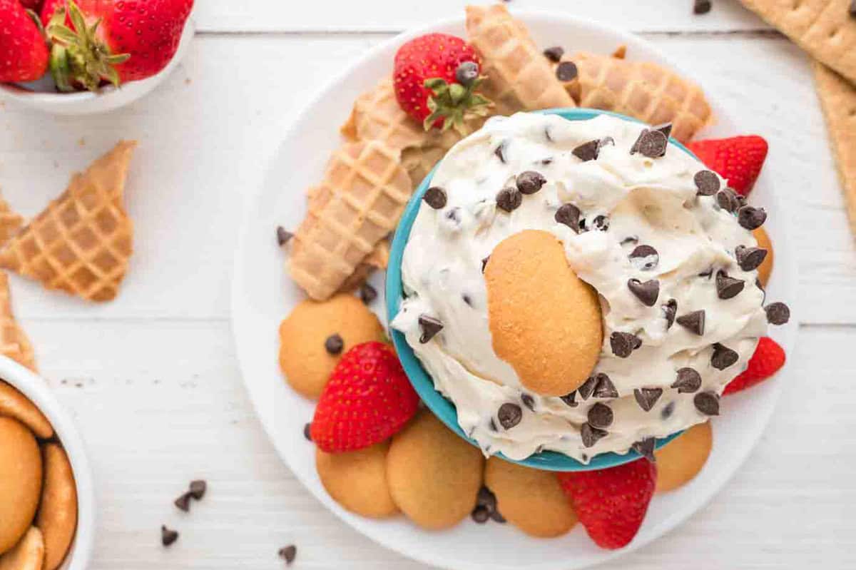 A bowl of cream cheese dip topped with chocolate chips and a vanilla wafer, surrounded by strawberries, vanilla wafers, and waffle cone pieces on a white plate.