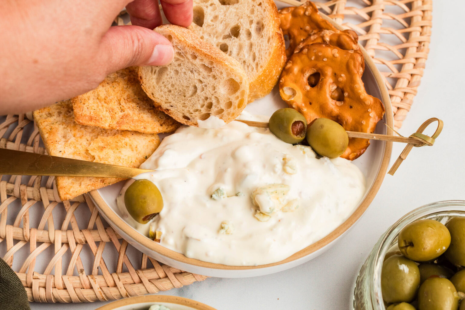 A hand dips a slice of bread into a plate with creamy dip, olives on a skewer, crackers, and pretzel chips on a woven placemat.
