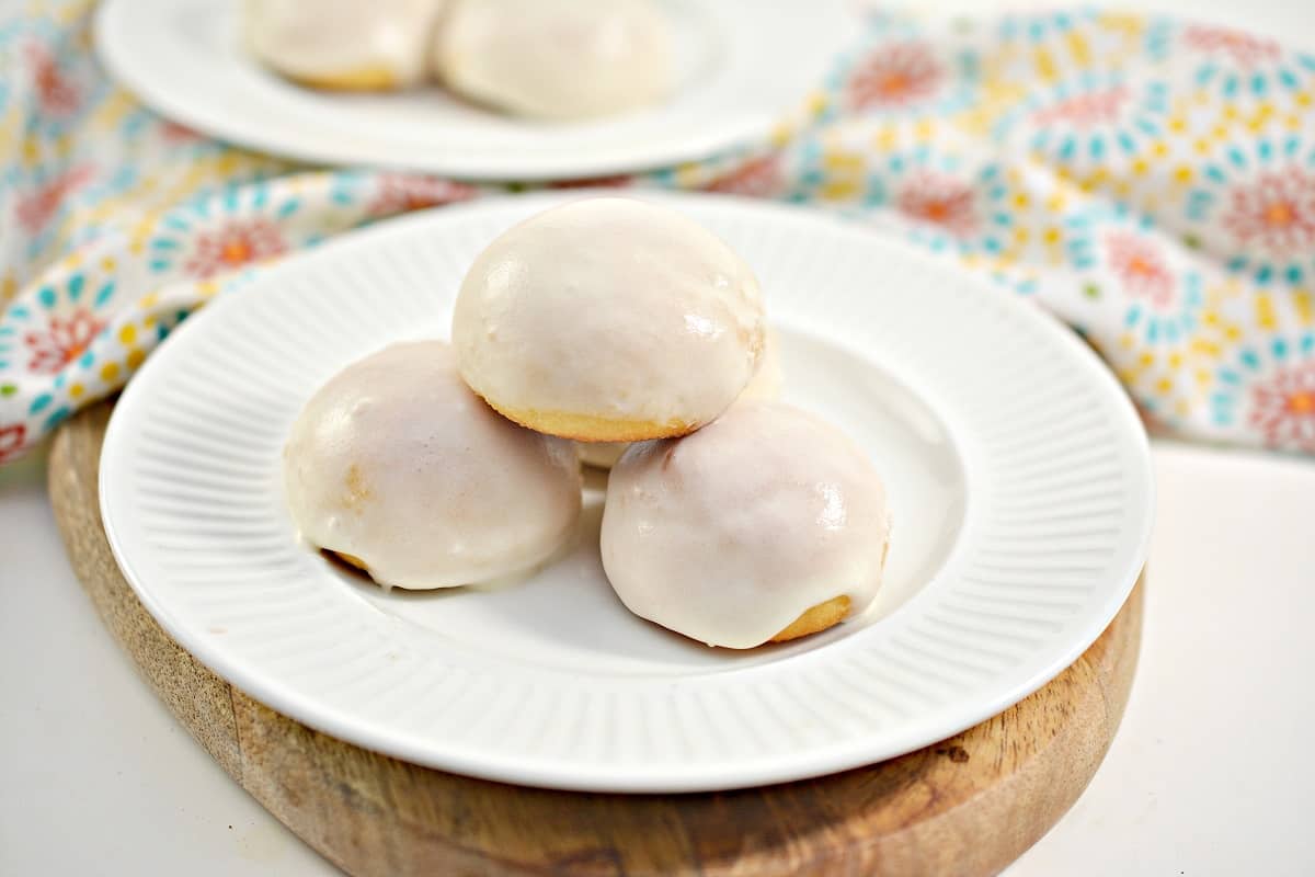 Three round, white-glazed cookies are arranged on a white plate set on a wooden board, with a patterned cloth in the background.