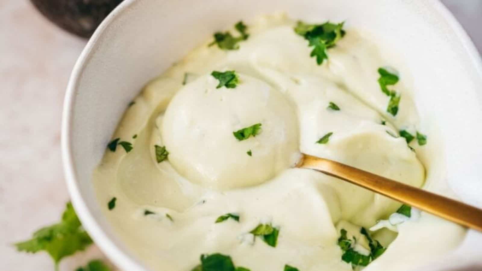 A bowl of creamy white sauce garnished with chopped green herbs, with a gold spoon resting inside.
