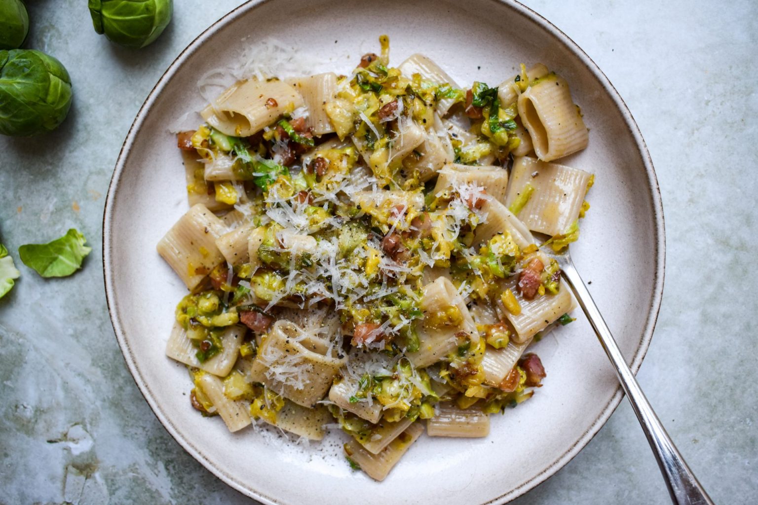 A plate of rigatoni pasta topped with a sauce containing chopped Brussels sprouts, bacon, herbs, and grated cheese, with a fork on the side.