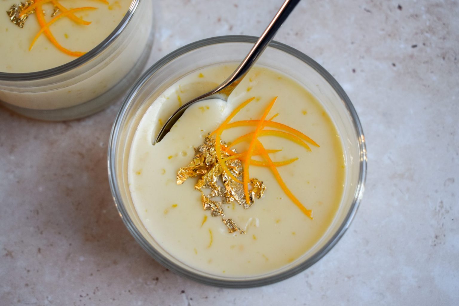 Glass cup of creamy dessert topped with orange zest and gold leaf, with a spoon resting inside. Another cup is partially visible in the background.