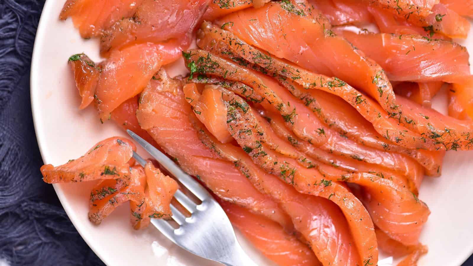 Thin slices of cured salmon garnished with herbs are arranged on a plate with a fork resting beside them.