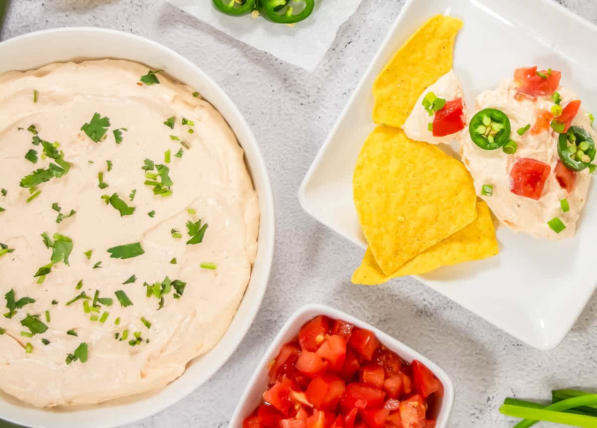 A bowl of creamy dip garnished with chopped herbs, served with tortilla chips, diced tomatoes, and sliced jalapeรฑos on the side.
