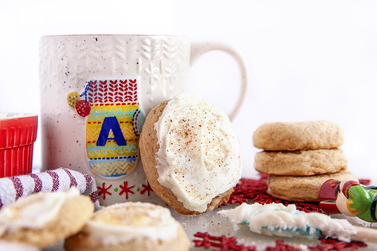 A frosted sugar cookie leans against a white patterned mug, with a stack of cookies, a red ramekin, and festive decor in the background.