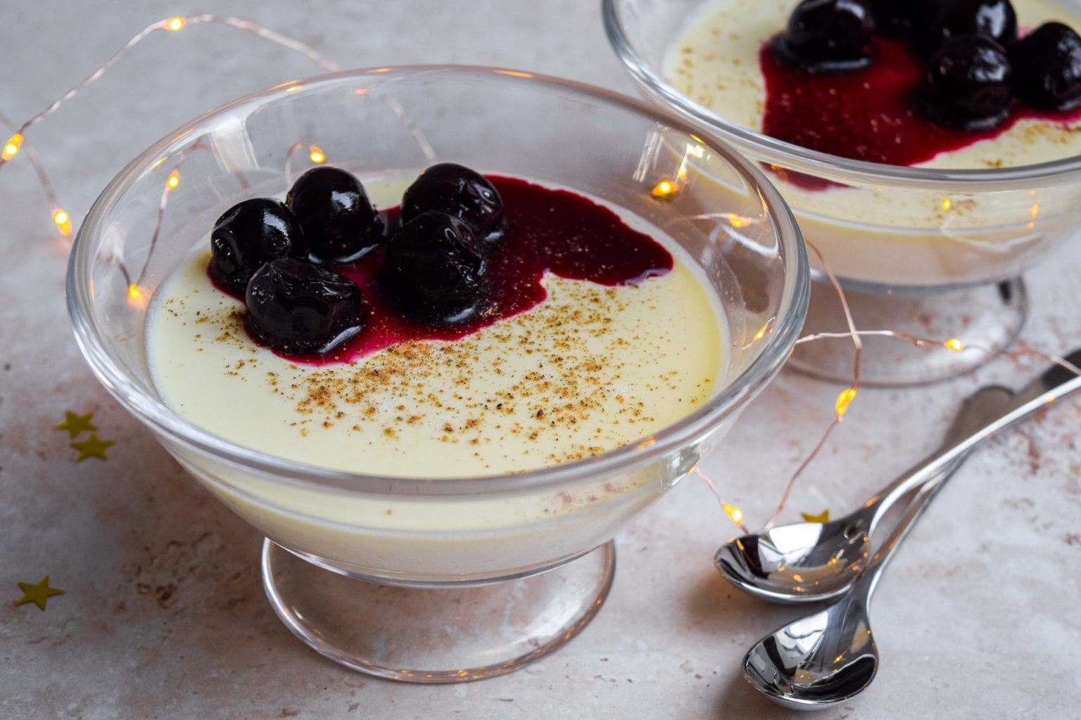 Two glass bowls of creamy dessert topped with dark berries and red sauce, lightly sprinkled with spice, next to two spoons and string lights on a light surface.