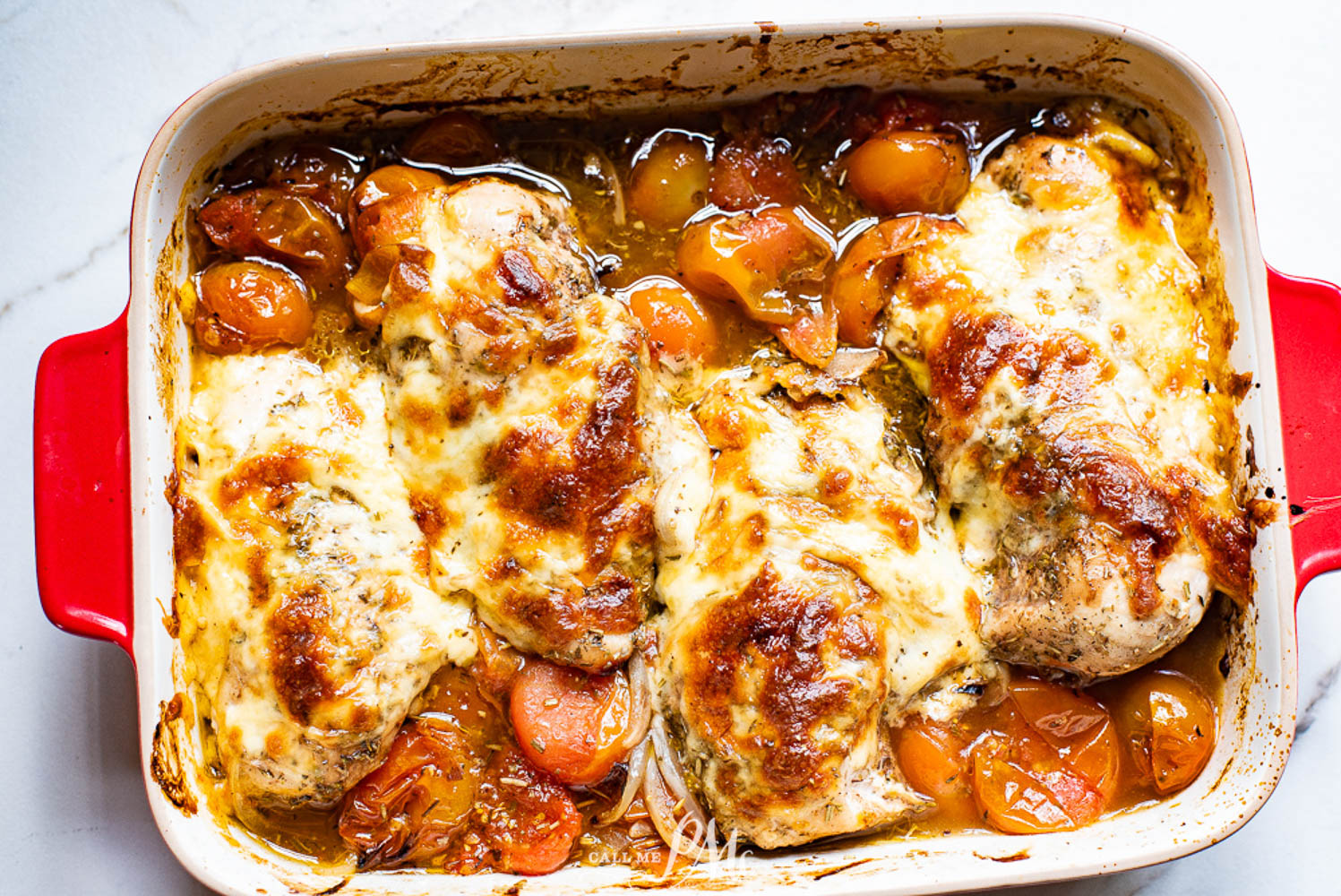 Baked chicken breasts topped with melted cheese and herbs, surrounded by cooked cherry tomatoes and sauce in a red casserole dish.