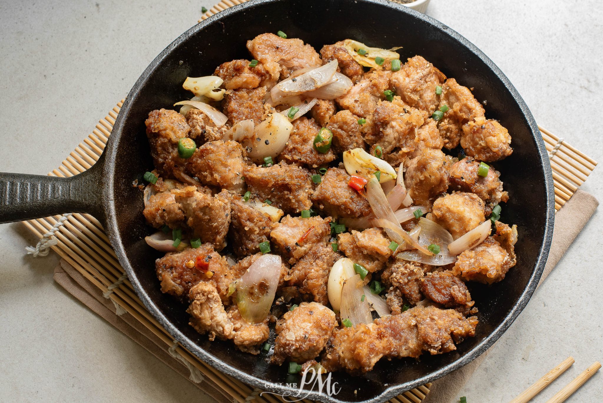 A cast iron skillet filled with fried chicken pieces, garnished with sliced onions, green herbs, and red chili, placed on a bamboo mat.