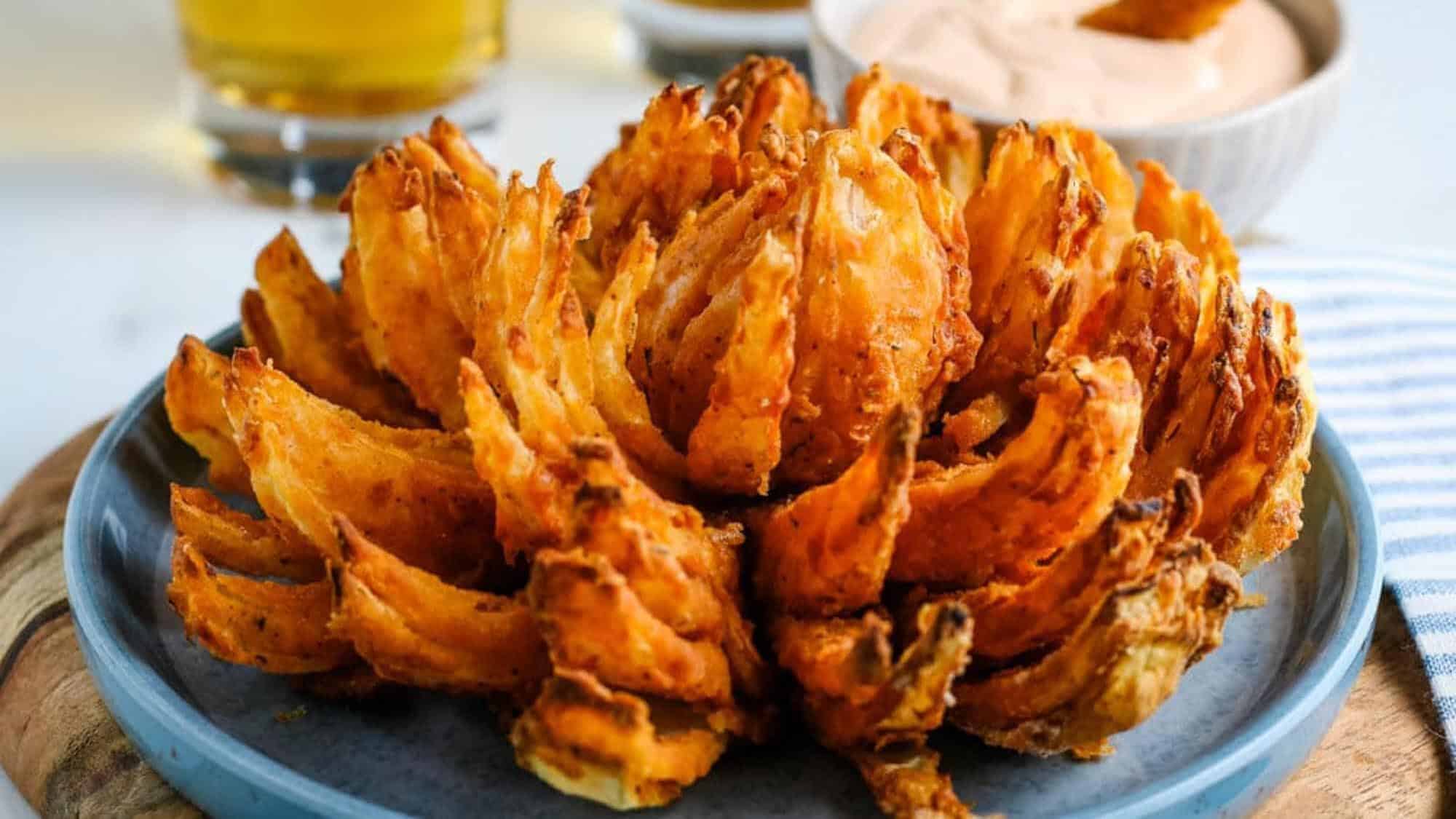 A fried blooming onion served on a blue plate with dipping sauce and a glass of beverage in the background.