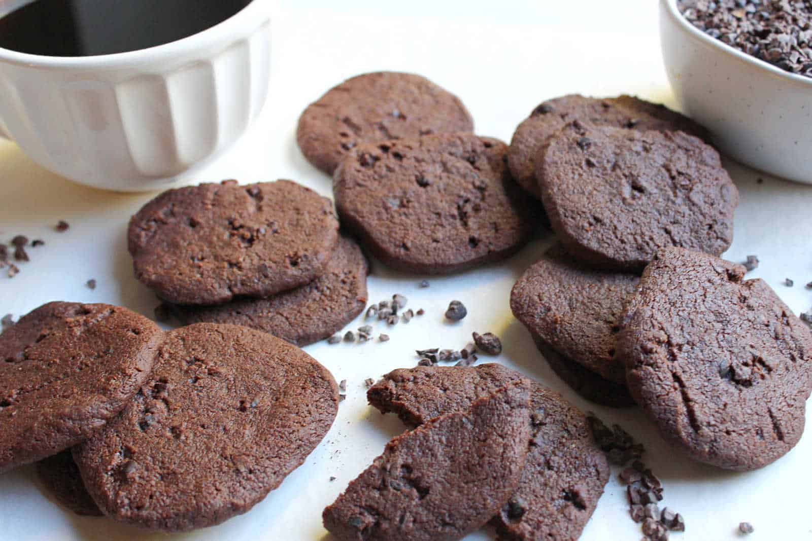 Several chocolate cookies are arranged on a white surface, with cocoa nibs scattered around and bowls containing a dark liquid and more nibs nearby.