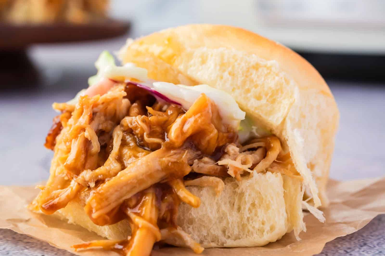 A pulled chicken sandwich with barbecue sauce and coleslaw on a soft bun, placed on brown parchment paper.