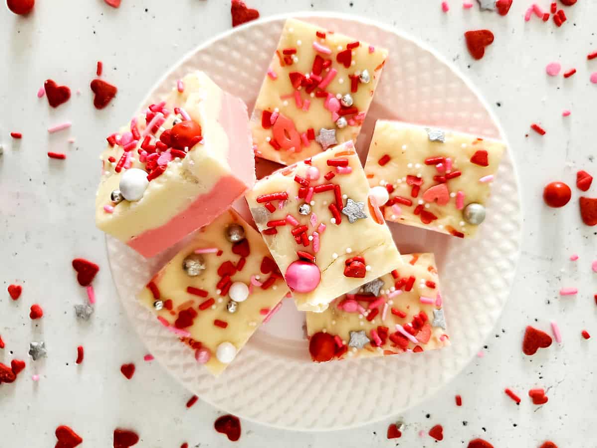 A white plate with several pieces of pink and white fudge topped with red, pink, and silver sprinkles and candy, on a white surface with heart-shaped decorations.