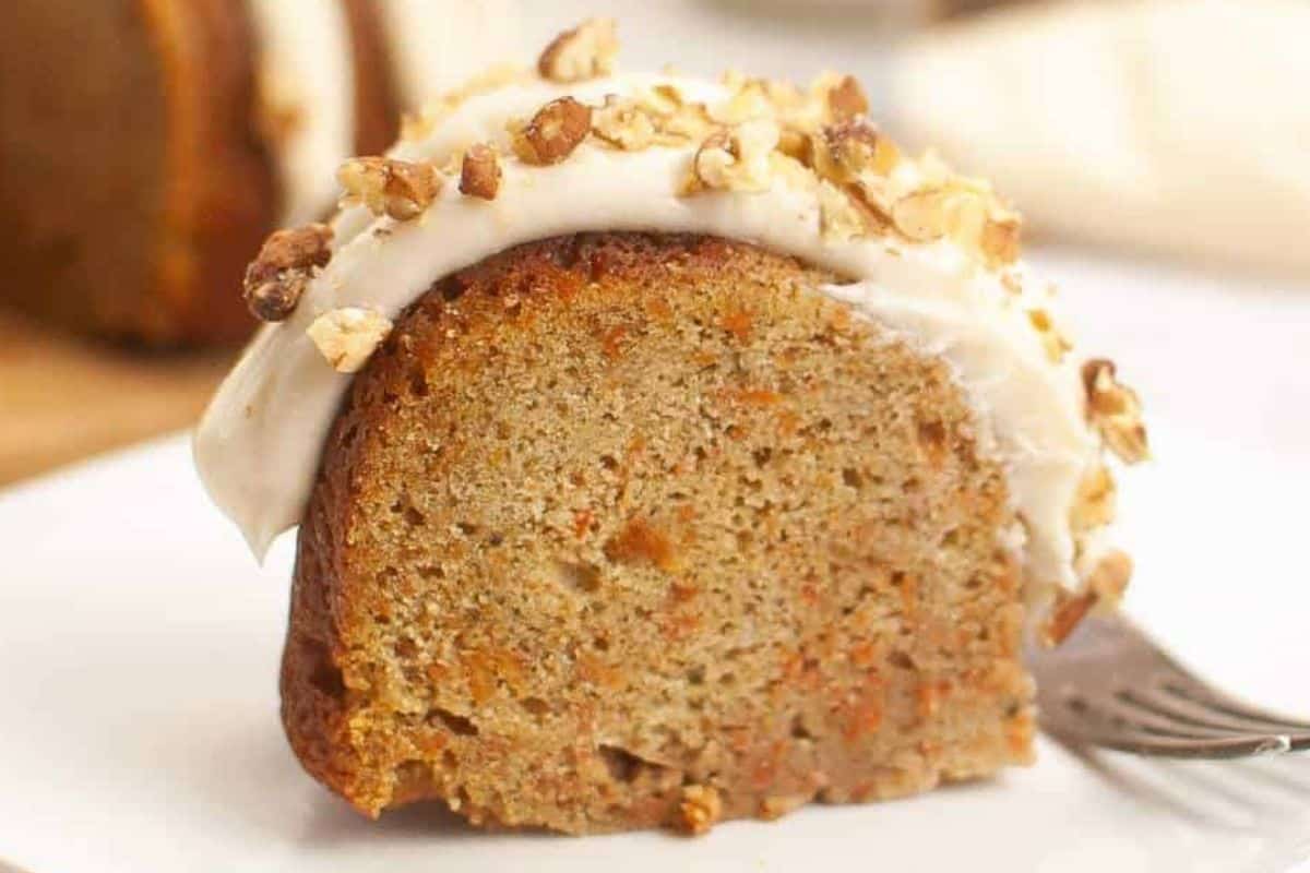 A slice of carrot cake topped with cream cheese frosting and chopped walnuts sits on a white plate with a fork beside it.