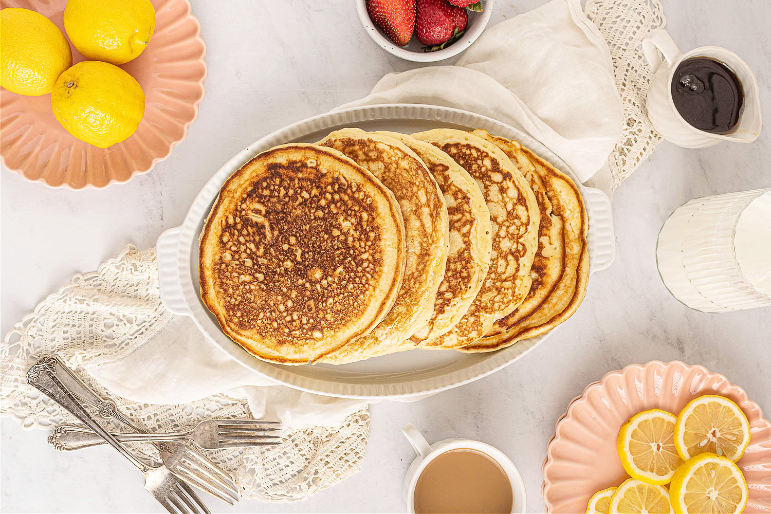 A white dish filled with pancakes sits on a table with plates of lemons, strawberries, a cup of coffee, forks, a jug of syrup, and a pitcher of milk.
