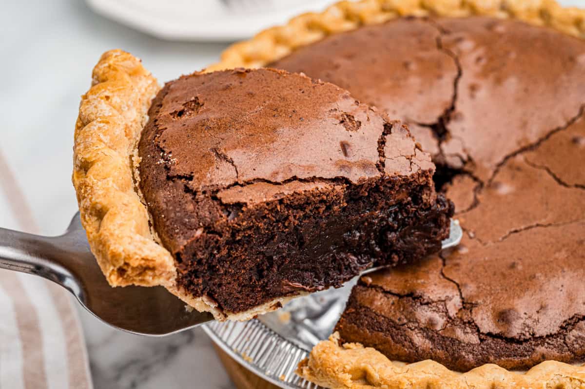A slice of chocolate pie with a cracked top and flaky crust is being lifted from a whole pie in a glass dish.