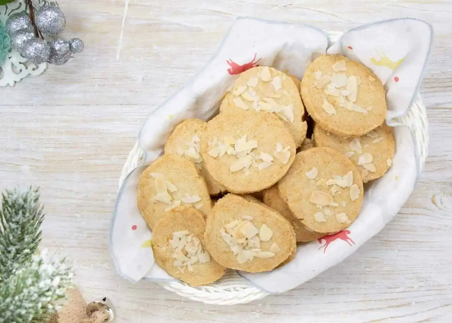 A basket lined with a white cloth holds several round cookies topped with almond flakes, placed on a light wooden surface.