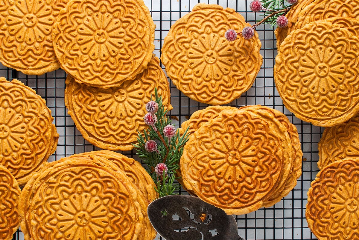 Decorative, round patterned cookies are arranged on a cooling rack with small sprigs of greenery and flowers, and a metal serving spatula is visible in the corner.