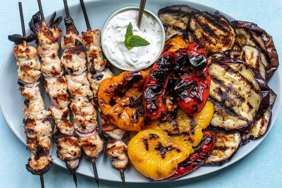 Grilled chicken skewers, charred bell peppers, and sliced grilled eggplant served with a bowl of creamy dip garnished with mint on a plate.