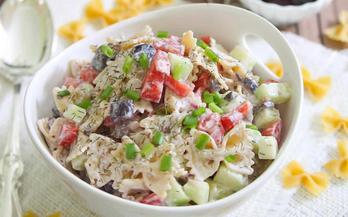A bowl of creamy pasta salad with bow tie pasta, diced cucumbers, tomatoes, black olives, green onions, and herbs.