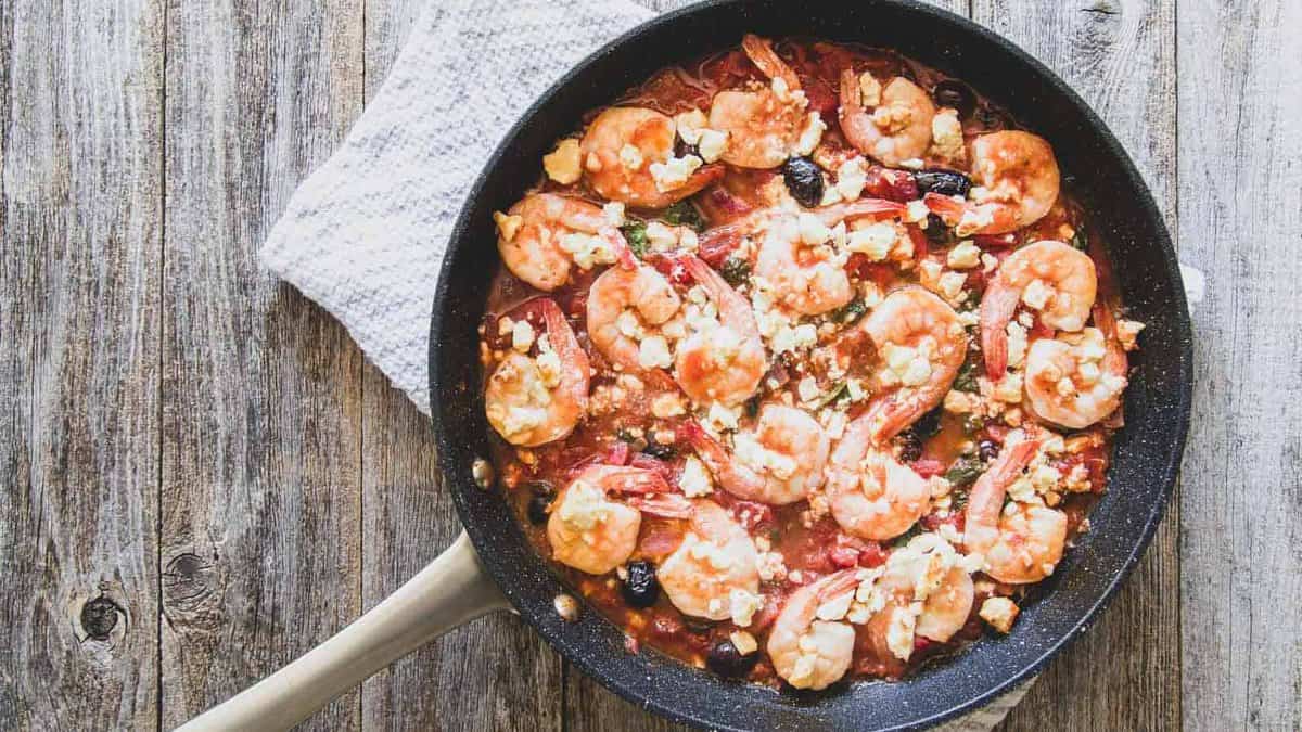 A skillet filled with shrimp, tomato sauce, olives, and crumbled cheese sits on a wooden surface next to a textured cloth.