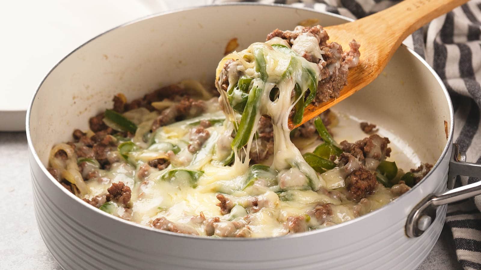 A skillet filled with cooked ground beef, sliced green bell peppers, onions, and melted cheese being stirred with a wooden spoon.