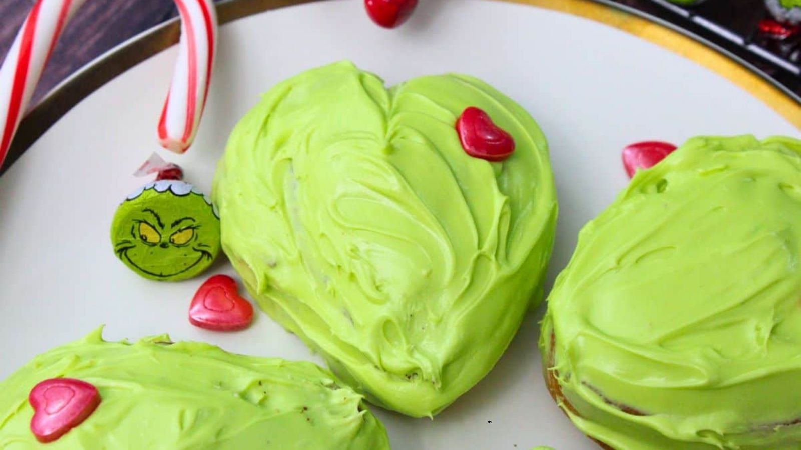 Heart-shaped cookie with green frosting and red heart candy on a plate, next to a candy cane ornament with a Grinch face.