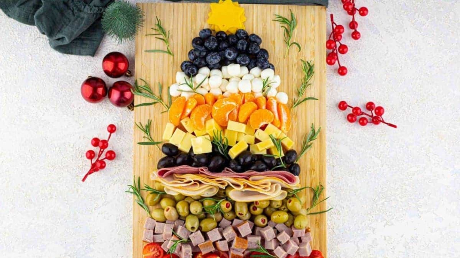 A wooden board arranged with fruits, cheese, meats, and olives in layers shaped like a Christmas tree, garnished with rosemary sprigs and festive decorations.