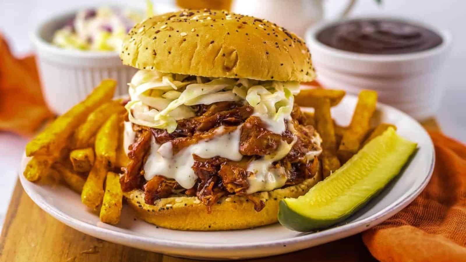 A pulled pork sandwich with coleslaw and sauce on a bun, served with French fries and a pickle spear on a white plate.