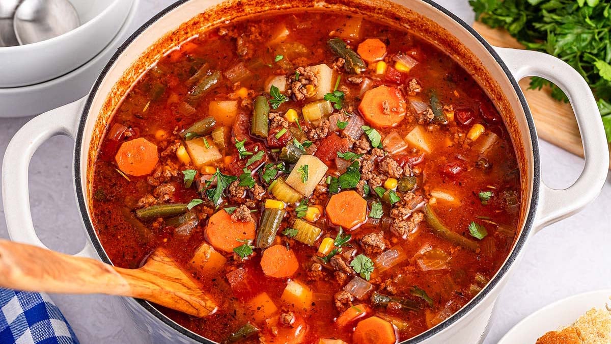A pot of vegetable beef soup with carrots, corn, green beans, potatoes, and ground beef, garnished with chopped parsley, sits on a countertop beside bowls and fresh herbs.