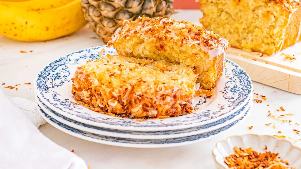 Two slices of toasted coconut bread are stacked on a decorative plate, with pineapples and bananas in the background.