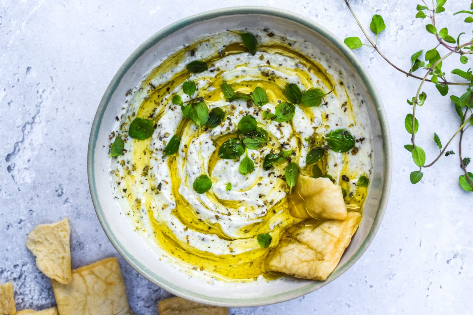 A bowl of creamy dip topped with olive oil and fresh herbs, served with pieces of pita bread on a light surface.