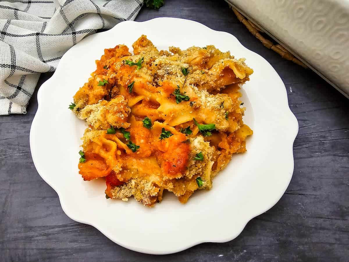 A serving of baked pasta topped with breadcrumbs and herbs is presented on a white plate, placed on a dark surface next to a checkered cloth.