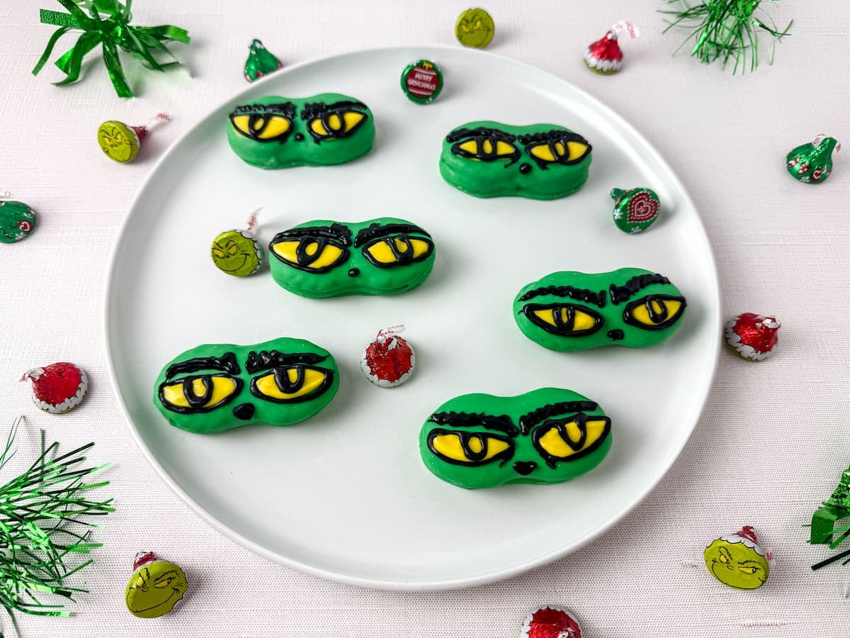 A white plate with six green cookies decorated to look like cartoonish yellow eyes with black outlines. Festive candy and green tinsel surround the plate on a white surface.