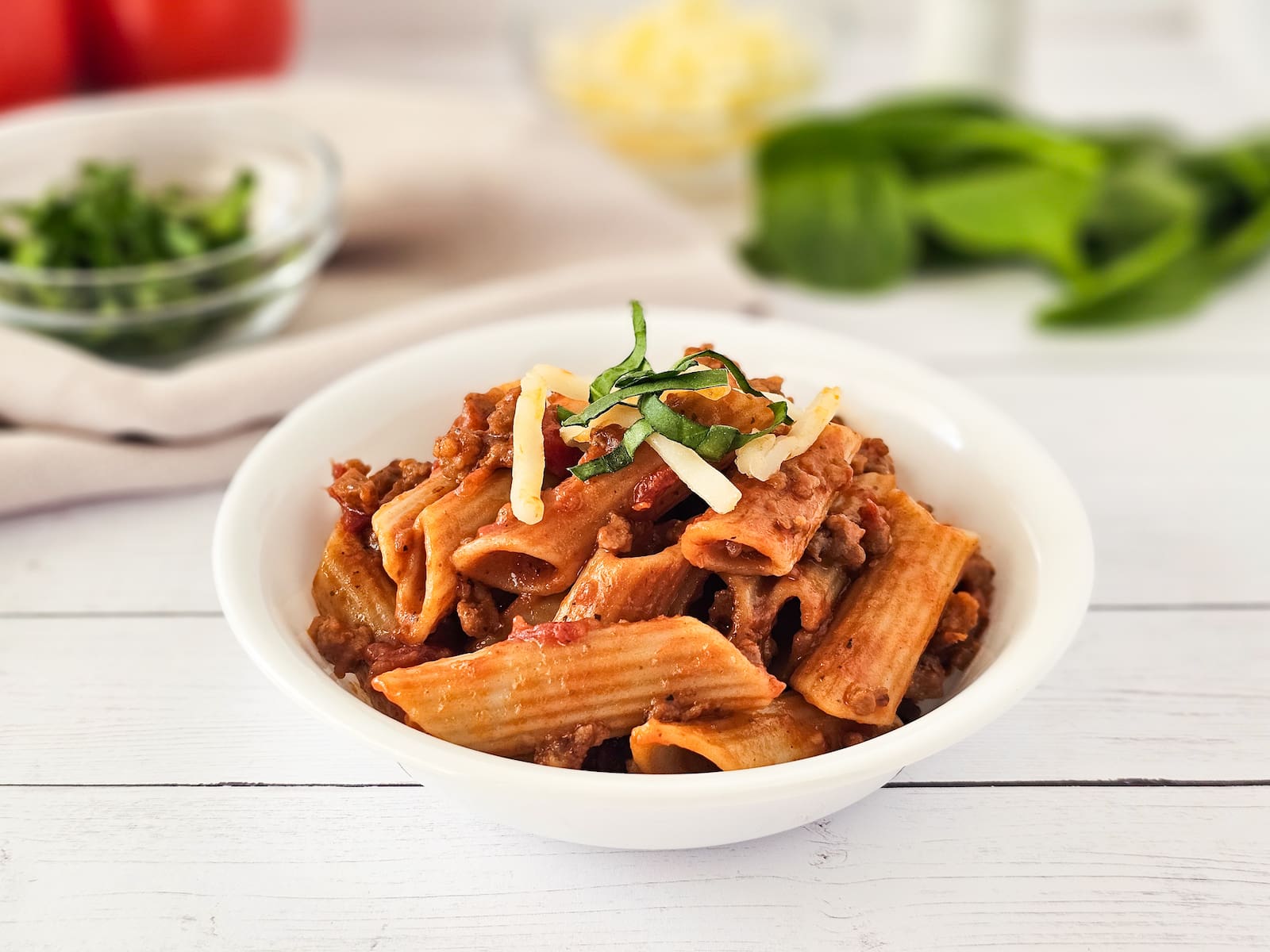A white bowl of rigatoni pasta with meat sauce, garnished with shredded cheese and basil, sits on a white wooden table with ingredients in the background.
