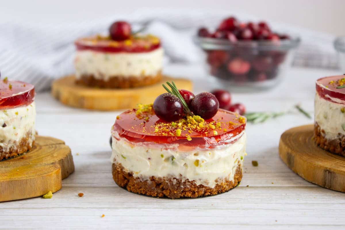 Mini cheesecakes with a crumb crust, creamy filling, and red berry jelly topping, garnished with berries and crushed pistachios, are displayed on wooden coasters.