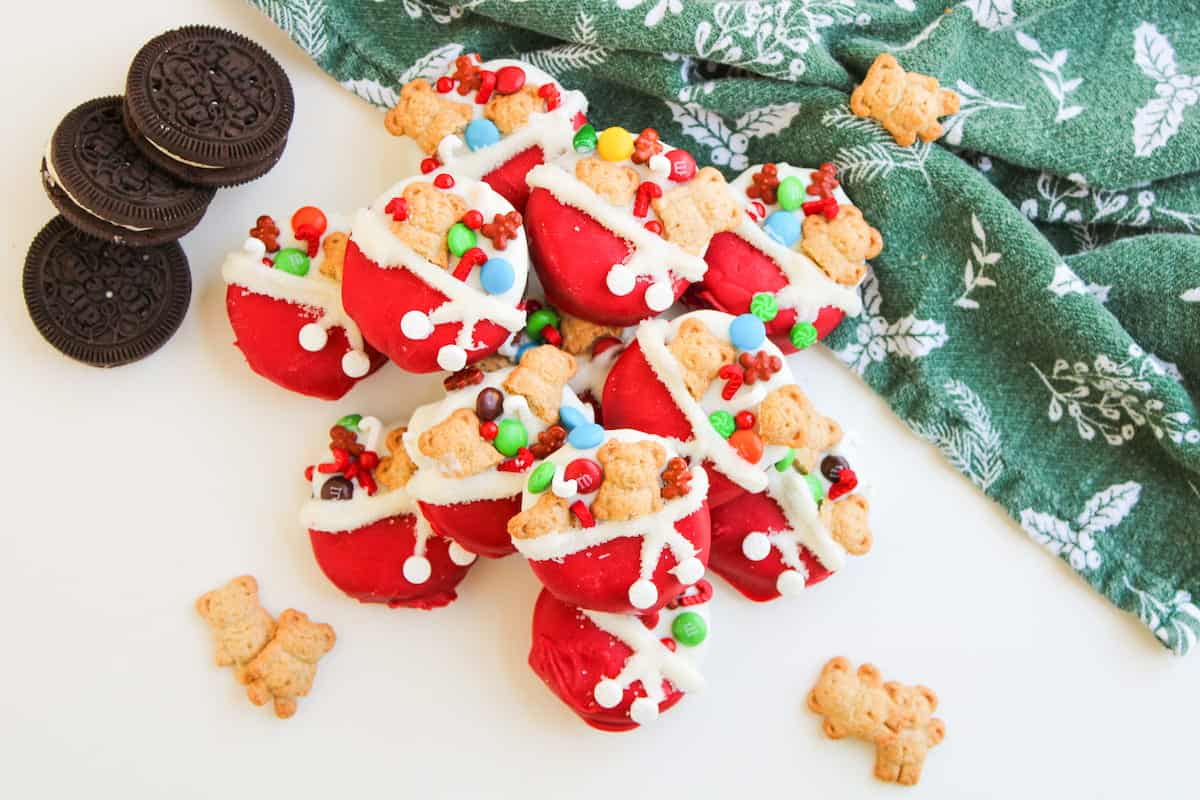 A pile of festive red-dipped Oreo cookies decorated with icing, candy pieces, and bear-shaped crackers on a white surface next to a green patterned cloth.